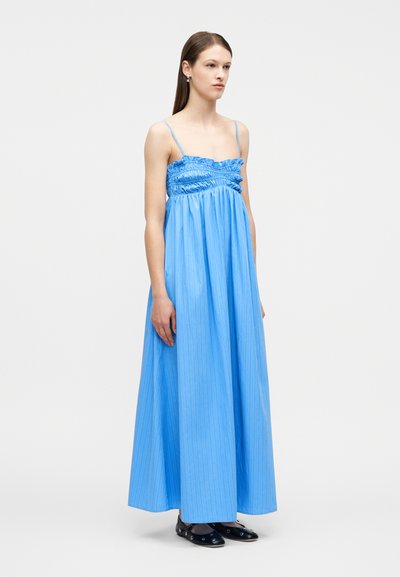 Γυναίκα που στέκεται με ένα μακρύ φόρεμα σε ανοιχτό μπλε χρώμα, χωρίς μανίκια, με ραφές στον κορσέ και λεπτές τιράντες, φορώντας μαύρα μπαλαρίνες με διακοσμητικές τρύπες.