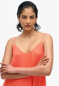 Koronkowa sukienka bez rękawów w kolorze koralowym z dekoltem w kształcie V, z plisowanym przodem i miękką teksturą. Ramiączka są cienkie, a materiał delikatnie opada na ciało.