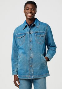 Chaqueta de mezclilla azul con cierre de botones en la parte delantera, dos bolsillos en el pecho y bolsillos laterales. Presenta costuras en contraste y diseño de cuello clásico.