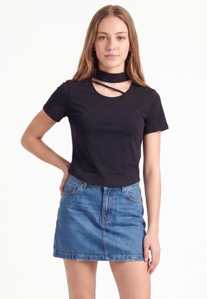 Jeune femme aux longs cheveux portant un haut noir à manches courtes avec col choker et découpe, associé à une jupe en denim bleu, debout avec une main sur la hanche.