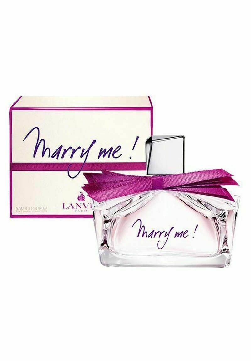 Genomskinlig parfymflaska med ett silverfärgat fyrkantigt lock, prydd med ett lila band. Etiketten visar "Marry me!" i lila skrivstil. Lådan har en liknande design.