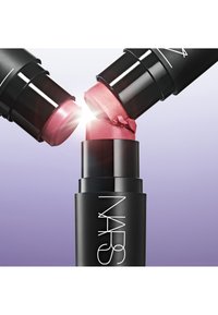 Deux tubes de rouge à lèvres NARS s'entrecroisent, l'un en noir mat avec un capuchon brillant, présentant des teintes roses et roses scintillantes, sur un fond dégradé violet.
