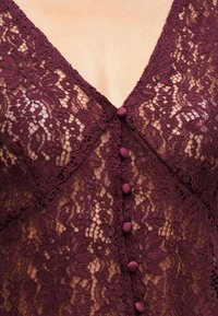 Peça em renda bordô com decote em V, apresentando padrões florais detalhados e botões decorativos revestidos de tecido na parte da frente.