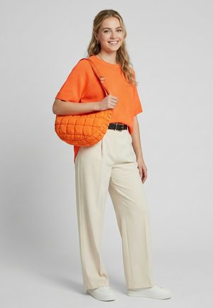 Smiling woman wearing an orange short-sleeve sweater and beige pants, holding a quilted orange shoulder bag, standing on a gray background.