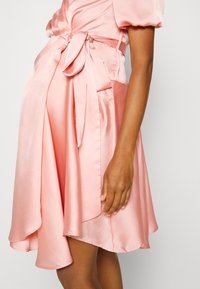 Vestido de cetim rosa com mangas curtas em forma de puff, cintura atada e uma bainha assimétrica, exibindo uma textura suave e uma silhueta fluida.