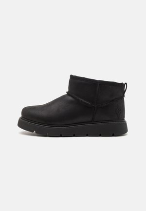 Botas negras hasta el tobillo, hechas de material liso con una puntera redondeada, detalles de costura y una suela de goma texturizada. Cuenta con una tira en la parte trasera para facilitar el calzado.
