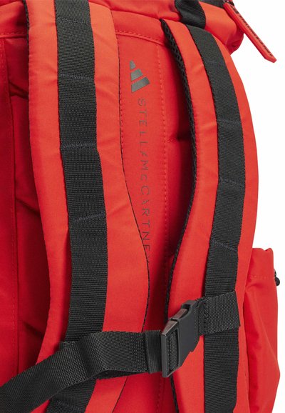 Red fabric backpack with black straps and hardware, featuring "STELLA McCARTNEY" branding and a side pocket for additional storage.