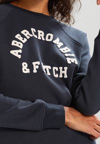Sudadera azul marino con cuello redondo, que presenta el texto bordado en blanco "ABERCROMBIE & FITCH" en el pecho; tejido suave y texturizado.