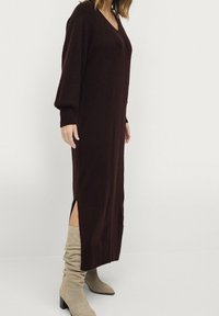 Lang, aansluitend jurk in donker bordeaux tricot met een V-hals, lange mouwen en zijsplitten. Gepaarde met lichtgrijze kniehoge laarzen.