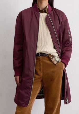 Personne portant une longue veste bomber bordeaux sur un pull en maille crème, une chemise à carreaux et un pantalon en velours côtelé marron, la main dans la poche.