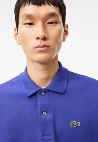 Pikétröja i starkblått med en strukturerad tyg, treknappslå och en liten grön krokodillogo på vänster bröst.