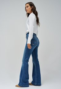 Jeans a zampa d'elefante blu con vita alta, caratterizzati da un classico design a cinque tasche e cuciture a contrasto. Abbinati a una camicia bianca a maniche lunghe.
