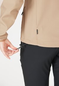 Beige lichtgewicht jack met zwarte ritssluitingen, verstelbaar trekkoord aan de onderkant, gedragen over zwarte broek, met een zijaanzicht van het kledingstuk.