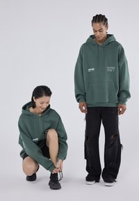 Πράσινα φαρδιά φούτερ με μπροστινές τσέπες και λευκό γραφικό κείμενο, συνδυασμένα με μαύρα τζιν με φθορές και μαύρα αθλητικά παπούτσια.