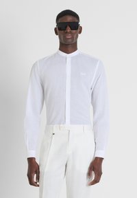 Camicia bianca con colletto rialzato, maniche lunghe e una texture sottile. Abbinata a pantaloni chiari e occhiali da sole oversize scuri.