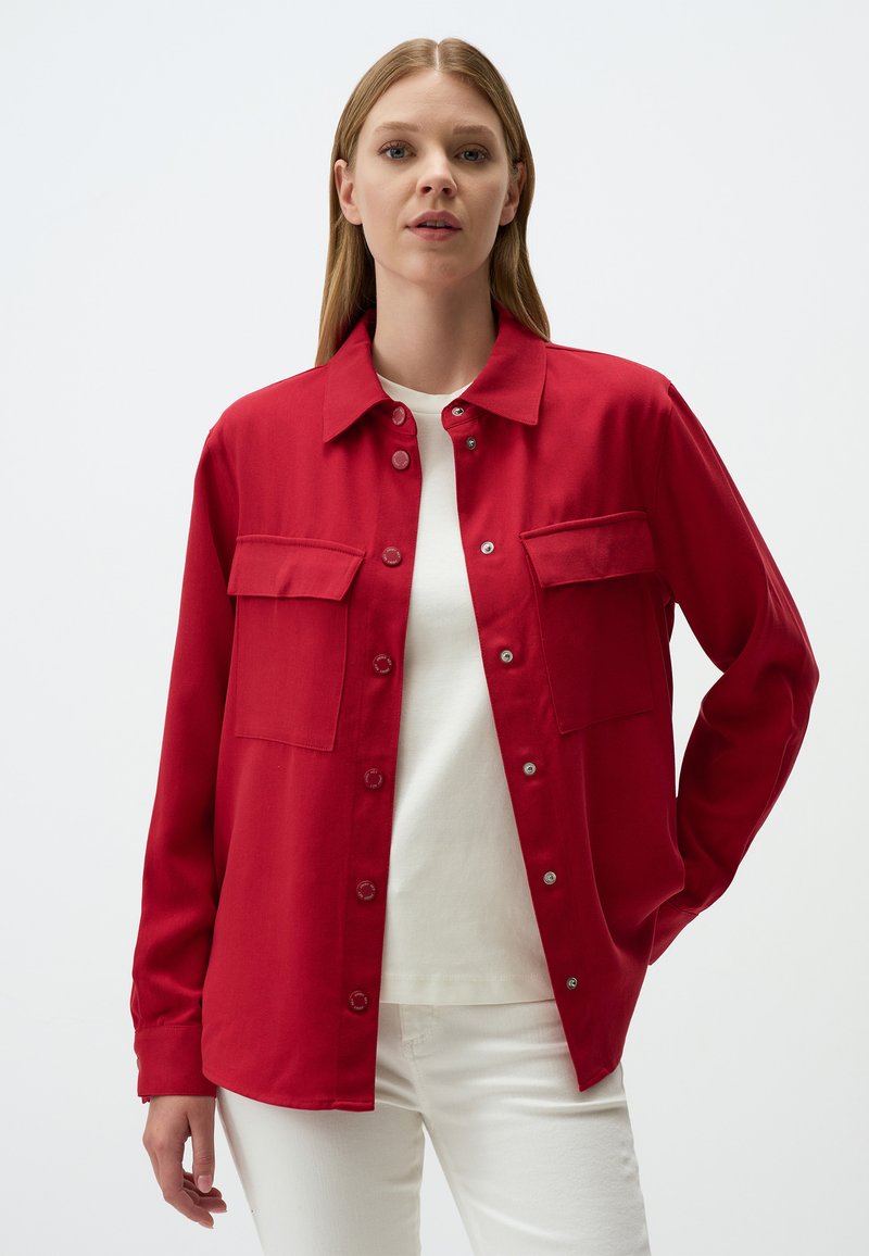 Veste rouge en tissu robuste. Présente un col, deux poches avant et des boutons-pression. Portée sur un haut blanc.