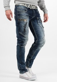 Jeans in denim di un blu scuro sbiadito con cuciture a contrasto, numerose tasche con zip e un marchio impresso lungo il lato.
