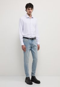 Camicia bianca a maniche lunghe, jeans slim-fit blu chiaro e stivali neri alla caviglia su un modello in piedi, con uno sfondo neutro e semplice.