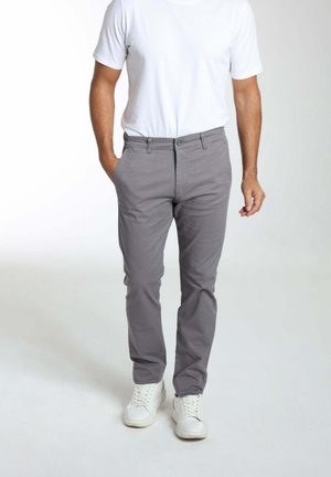 Homme portant un pantalon gris, un t-shirt blanc et des baskets blanches, debout avec une main dans la poche sur un fond clair.