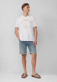 T-shirt en coton blanc avec un imprimé graphique, associée à un short en denim bleu clair et des sandales à brides marron avec une boucle décorative.
