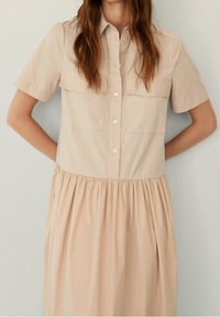 Abito beige con bottoni, maniche corte, due tasche sul petto, gonna plissettata e tessuto di cotone liscia.