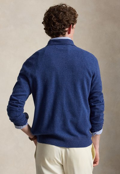 Άνδρας με καστανά σγουρά μαλλιά φορά μπλε πουλόβερ πάνω από ριγέ πουκάμισο και μπεζ παντελόνι, στέκεται γυρισμένος με τα χέρια στη μέση.