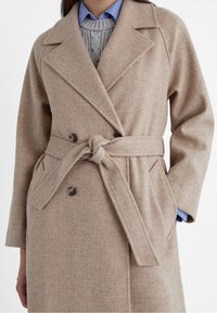 Beżowy płaszcz z wełny z podwójnym zapięciem, z paskiem w talii i klapami. Posiada dwa guziki i boczne kieszenie dla funkcjonalności.