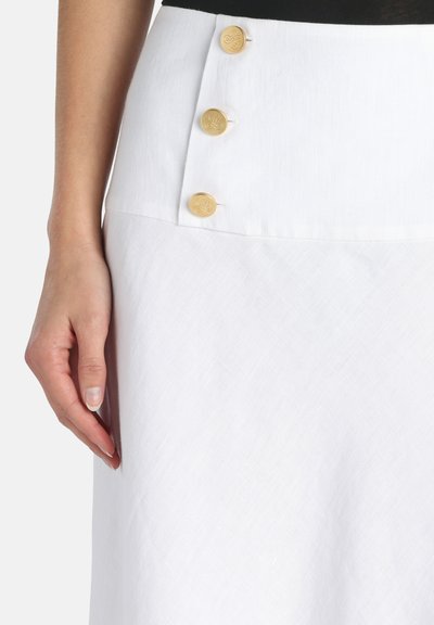 Nærbillede af et hvidt skørt med tre guldknapper på siden og en hånd hvilende ved siden af skørtet.