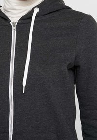 Tmavě šedá mikina na zip s bílými šňůrkami a stříbrným viditelným zipem, nošená přes bílý rolák.