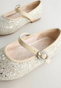 Tüdrukute sädelevad beežid Mary Jane kingad lillemustritega sisetaldadega ning rhinestone'idega kaunistatud ümmarguste lukutustega neutraalsel taustal.