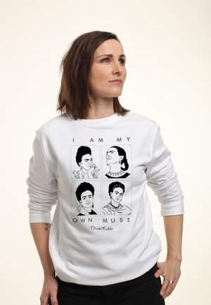 Kvinde med skulderlangt brunt hår iført en hvid sweatshirt med fire sort-hvide skitser af Frida Kahlo og teksten "Jeg er min egen muse."