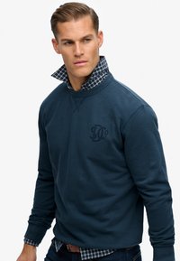 Granatowy sweatshirt z widocznym logo, z plaidowym kołnierzem pod spodem. Tkanina wydaje się miękka, z prążkowanymi mankietami i dekoltem.