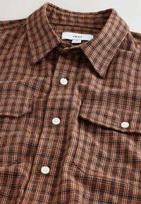 Braun und schwarz kariertes Flanellhemd mit einem Knopfkragen, zwei Brusttaschen und weißen Kunststoffknöpfen. Weiche Textur.