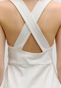 Witte jurk met een kruislingse rugontwerp, vervaardigd uit zachte stof en een getailleerde pasvorm. De rug is blootgesteld en heeft een discreet ritsje.