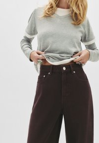 Siv sveter s dolgimi rokavi in V-izrezom, preplastljen preko belega topa, skupaj z visokim pasom temno rjavih širokih hlač, ki vključujejo sprednje žepe in gumb.