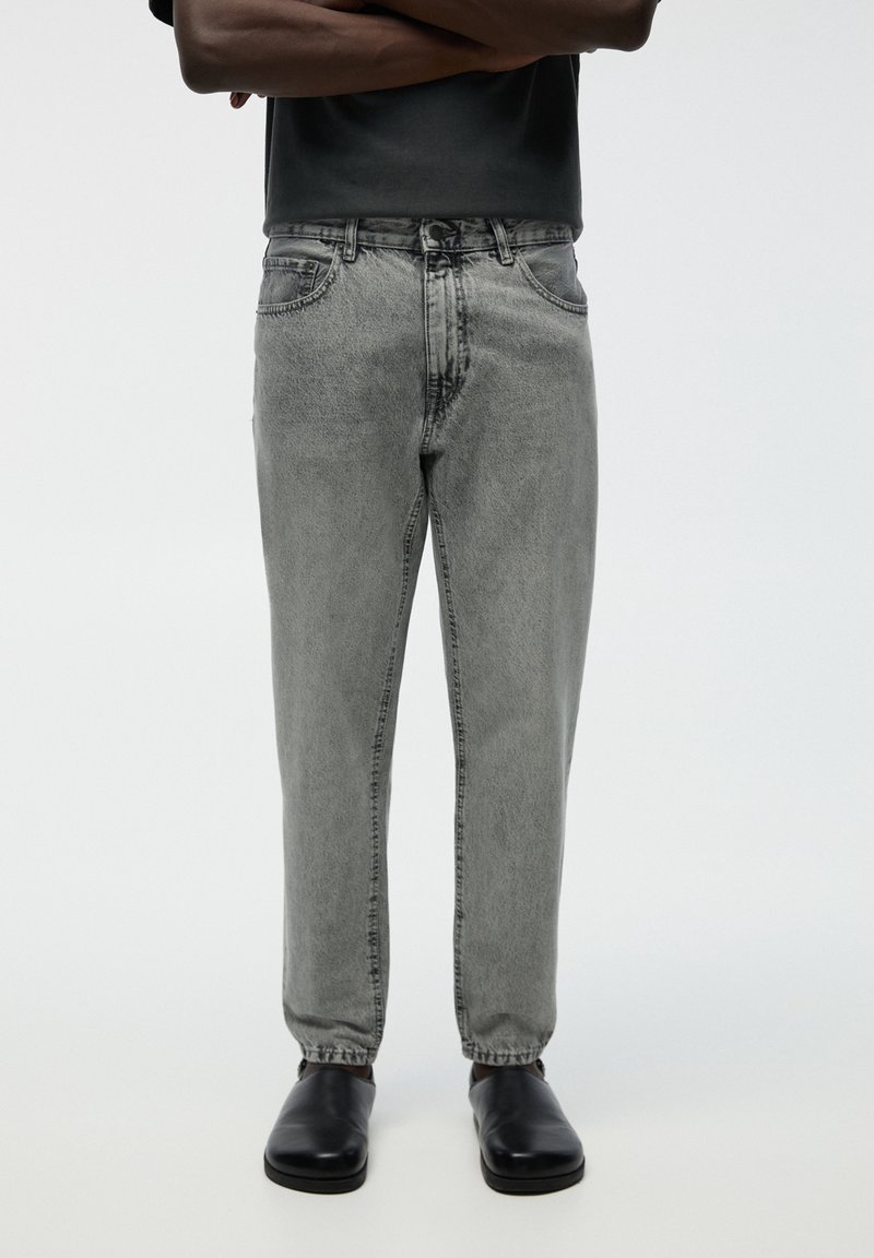 Personne portant un jean gris délavé taille haute, des chaussures noires et une chemise noire à manches courtes, les bras croisés, debout devant un fond blanc.