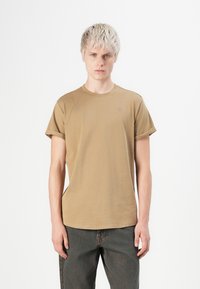 Ung mann med kort blond hår, iført en beiget T-skjorte med oppbrettede ermer og mørk denimjeans, som står mot en hvit bakgrunn.