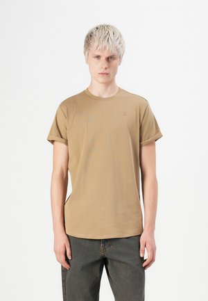 Joven con cabello rubio corto que lleva una camiseta beige de manga arremangada y jeans oscuros, de pie contra un fondo blanco.