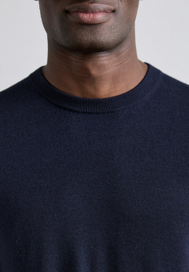Suéter azul marino de cuello redondo hecho de tejido de punto suave y texturizado. Cuenta con un cuello acanalado y una confección sin costuras, mostrando un diseño minimalista.