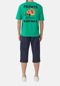 T-shirt en coton vert avec du texte noir indiquant "SURFWAX au goût de PAPAYE" et un graphique de fruit de papaye, assorti à un short cargo bleu marine.