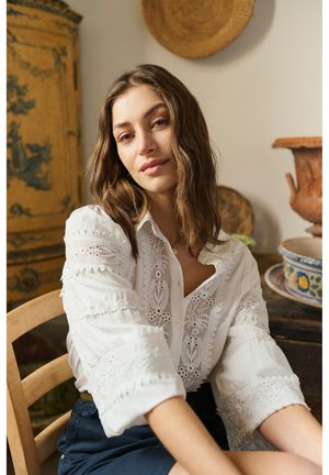 Tânără cu păr castaniu ondulat, purtând o bluză albă brodată, așezată pe un scaun de lemn într-o cameră rustică.