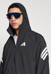 Zwarte windjack met kap, voorzien van witte strepen op de mouwen en een logo. Model draagt futuristische donkere zonnebril met reflecterende lenzen.