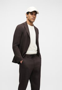 Homme portant un costume marron foncé avec un t-shirt blanc et une casquette beige clair, debout les mains dans les poches devant un fond uni.