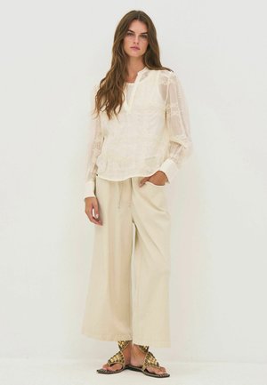 Femme aux longs cheveux bruns portant un chemisier en dentelle crème, un pantalon évasé et des sandales dorées cloutées, debout devant un fond blanc uni.