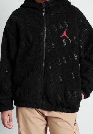 Persona che indossa una giacca Jordan nera zippata con logo Jumpman rosso e pantaloni beige con logo Jumpman nero visibile sulla coscia.