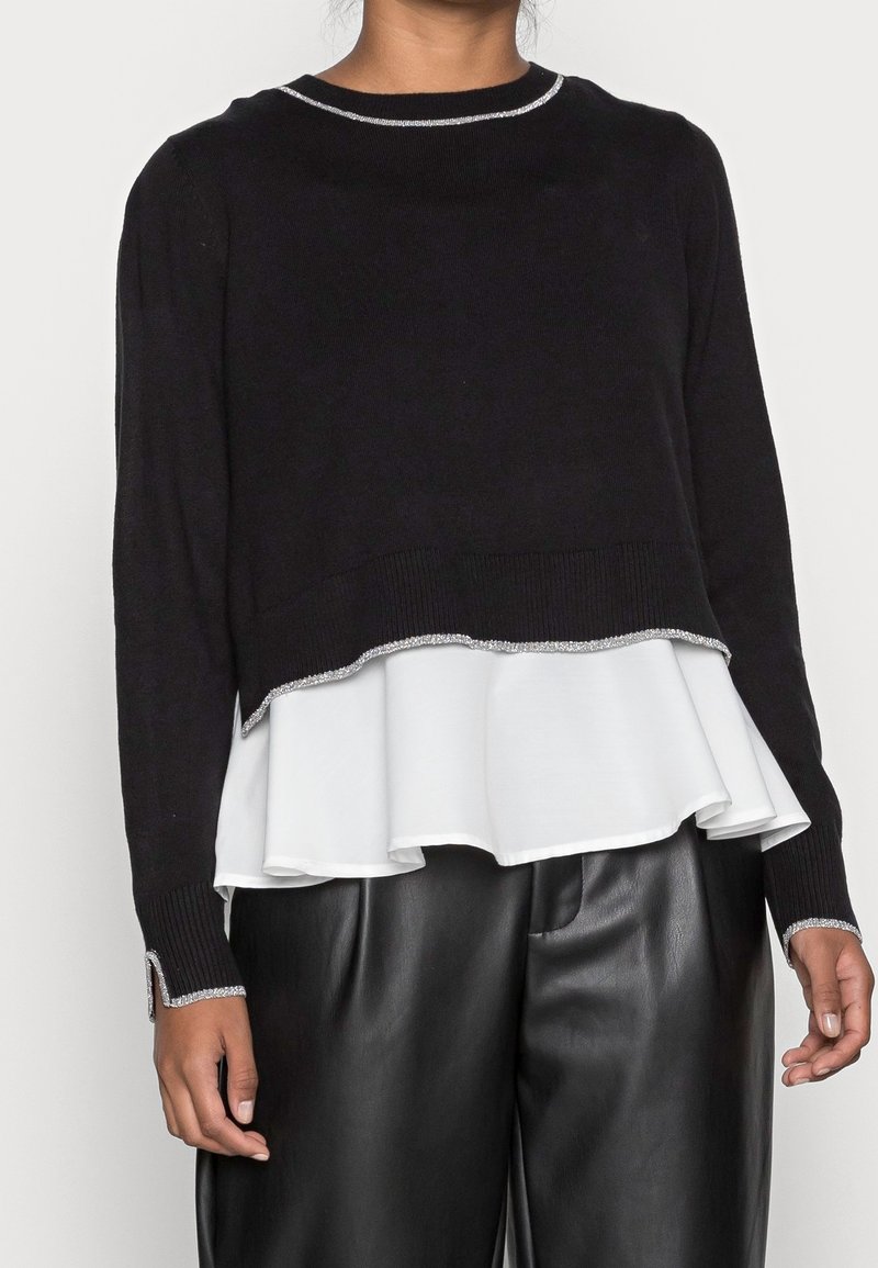Black knitted sweater with a layered white ruffle hem, silver trim along the neckline and cuffs, and ribbed texture, paired with black leather pants.