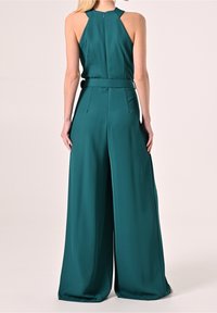 Mujer que lleva un mono sin mangas en tono aguamarina con pantalones de pierna ancha y escote halter, de pie de espaldas contra un fondo liso.