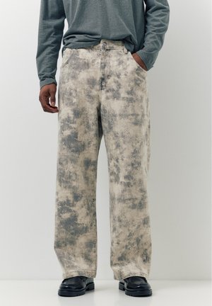 Férfi bővebb szabású nadrág szürke és bézs absztrakt mintával, fekete cipővel és begombolt, hosszú ujjú csíkos inggel párosítva.