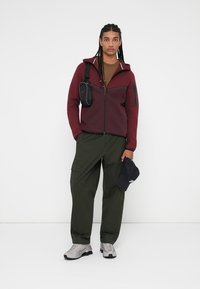 Casaco zip-up bordeaux com capuz e detalhes pretos, t-shirt castanha, calças cargo verdes, sapatilhas cinzas de desporto e um boné preto.