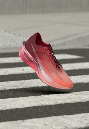 Raudona ir rožinė Nike bėgimo batelio su raišteliais ore virš zebro perėjos ant asfalto kelio.